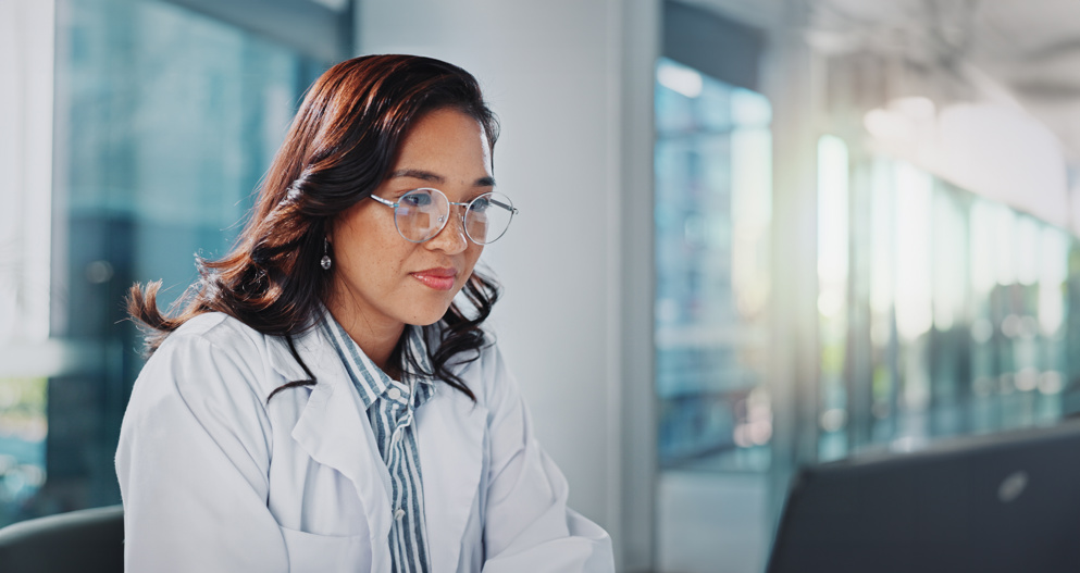 Healthcare professional wearing a lab coat seated in a bright clinic, looking at a computer screen
